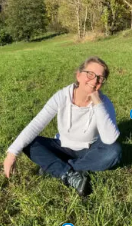 Woman sitting cross-legged on grass in a park, resting her head on her hand and smiling.