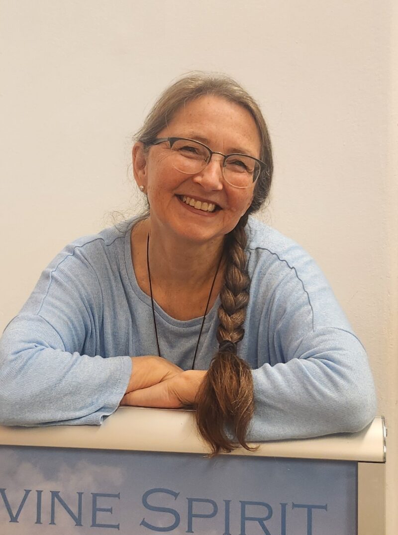 Smiling woman with glasses and a long braid, wearing a light blue sweater, resting arms on a table.