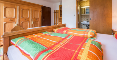 Bedroom with a wooden bed and a bright plaid quilt; a large wooden wardrobe and an open bathroom door are visible in the background.