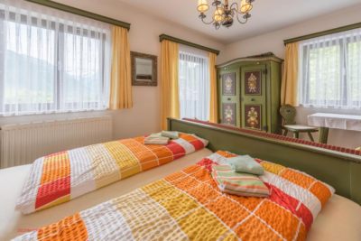 Twin beds in a sunlit room with colorful orange-yellow quilts, green furniture, and sheer-curtained windows.